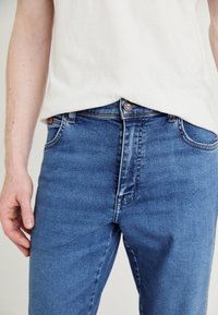 Kék farmernadrág, slim fit szabásban, közepes derékkal, enyhe kopással és standard zsebekkel, fém kiegészítőkkel a záráshoz.