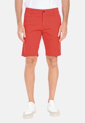 Uomo che indossa pantaloncini rossi vivaci fino al ginocchio, scarpe da ginnastica bianche e una camicia bianca, in piedi davanti a uno sfondo semplice.