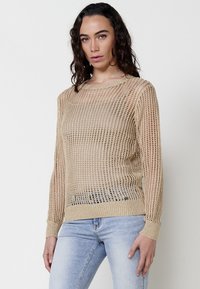 Beige kötött pulóver, hálós mintázattal, hosszú ujjal, bordázott mandzsettával és kerek nyakkivágással, világoskék farmerral párosítva.