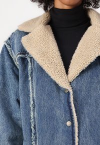 Veste en jean avec un col en fausse fourrure beige. Présente des bords effilochés et des boutons-pression en argent. Un pull à col roulé noir en dessous.