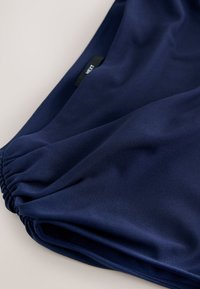 Tissu bleu marine avec bord élastiqué et petite étiquette noire affichant un texte blanc partiel "NEXT" sur un fond neutre.