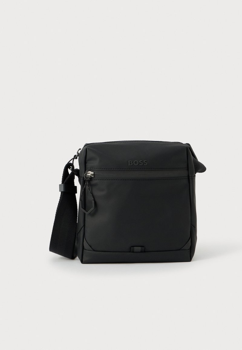 Borsa a tracolla nera in materiale liscio e opaco con tasca frontale con zip, dettaglio logo e tracolla regolabile. Forma rettangolare compatta.