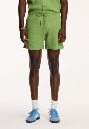 Groene shorts met een waffle-structuur en een trekkoord in de taille, gecombineerd met lichtblauwe sneakers en witte sokken, wat een casual, sportieve look laat zien.