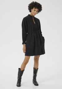 Vestido de mezclilla negra con mangas largas, collar con volantes, placket frontal y cinturón atado. Presenta grandes bolsillos laterales y un corte holgado.