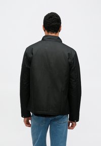 Jachetă neagră, ușoară, cu guler ridicat, croială dreaptă și manșete cu nasturi, asortată cu blugi albaștri. Design simplu, fără modele sau accente vizibile.