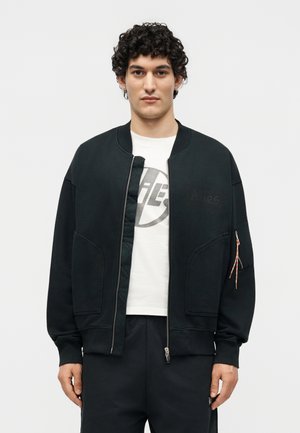 Jaunas vyras su garbanotais plaukais dėvi juodą bomber stiliaus striukę su užtrauktuku ant baltos marškinėlių su grafiniu piešiniu, stovėdamas prieš šviesią vientisą foną.