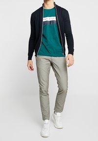 Marinblå zip-up cardigan över en turkos t-shirt med vita och grå ränder, i kombination med ljusgrå byxor och vita sneakers. Släta texturer genom hela outfiten.