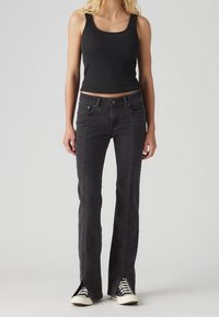 Débardeur noir côtelé associé à un jean évasé foncé avec des fentes latérales et un détail de couture texturée. Complété par des baskets noires et blanches.