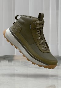 Olive roheline kõrge tõusuga sneaker tekstuuriga kangaga, kontrastsete valge ja kummitekkidega tallaga, paelte detailiga ja väljendunud pahkluu toetusega.