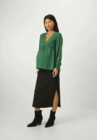 Blouse de maternité verte avec un col en V et un détail froncé, associée à une jupe midi noire avec une fente sur le côté. Bottines noires.