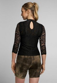 Schwarzes Spitzenoberteil mit Dreiviertelärmeln, gewellten Kanten und einem Schlüsselloch-Rückendetail, kombiniert mit braunen Shorts mit strukturierter Oberfläche.