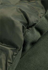 Tela exterior verde con una textura suave y brillante, y un forro de vellón texturizado y suave. Integración perfecta de materiales en un diseño en capas.