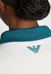 Λευκό πόλο πουκάμισο με υφή, με γαλάζιο ριμπ κολάρο και ένα στυλιζαρισμένο λογότυπο σε γαλάζιο στην πλάτη.