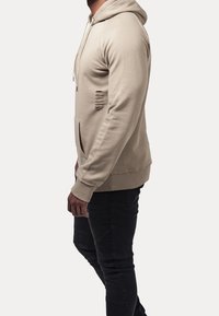 Beige hoodie featured with a front pocket, a drawstring hood, and distressed detailing on the left side. Paired with dark textured pants.