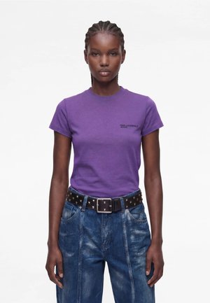 Frau mit geflochtenem Haar, die ein lila T-Shirt und blaue Jeans mit schwarzem Nietengürtel trägt, steht vor einem schlichten weißen Hintergrund.