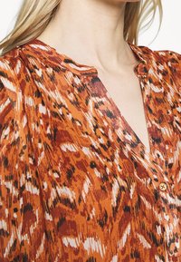 Gros plan d'une femme portant un chemisier imprimé animalier orange et marron avec un col en V et un seul bouton visible.