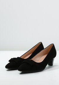 Une paire de chaussures pour femmes en suedine noire à petits talons avec des orteils pointus et de petits nœuds décoratifs à l'avant.