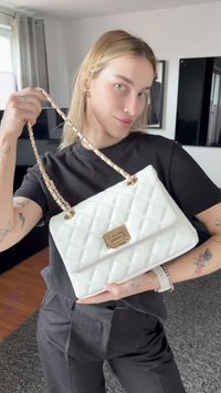 Világos hajú és tetoválásos személy fehér, kockás mintázatú kézitáskát tart, arany láncpánttal és kapoccsal egy modern nappaliban.