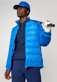 Homem a usar um boné azul, uma jaqueta puffer azul, uma polo azul-marinho e calças, segurando um taco de golfe sobre o ombro com uma luva branca na mão direita.