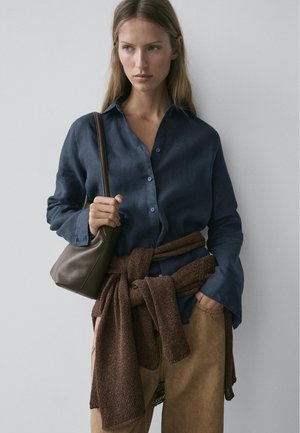 Ung kvinde iført mørkeblå knappet skjorte og brune bukser, der holder en mørkebrun taske med en brun sweater bundet omkring taljen.