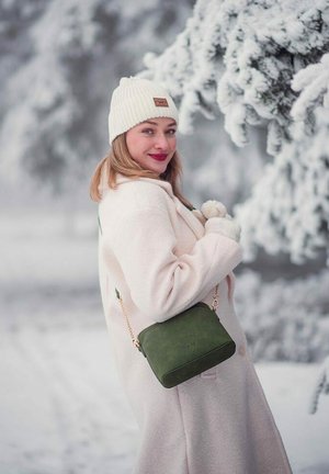 Frau in weißem Mantel und gestrickter Mütze lächelt und hält eine grüne Handtasche im Freien nahe schneebedeckter Kiefern in winterlicher Umgebung.