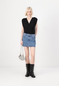 Top negro envolvente, minifalda de mezclilla azul y botas negras hasta la rodilla. La modelo lleva un pequeño bolso de malla plateada. Fondo minimalista.