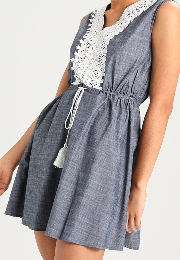 Robe sans manches bleue avec des détails en dentelle blanche et une taille à cordon, portée par une personne avec le bras détendu sur le côté.