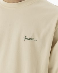 Bež majica od pamuka s okruglim izrezom i zelenim kurzivnim logom na lijevoj strani prsnog koša. Glatka tekstura, klasičan kroj.
