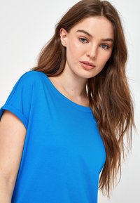 Blå kortærmet t-shirt lavet af blødt stof med en afslappet pasform og bred halsudskæring. Overfladen er glat, uden mønstre eller pynt.
