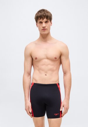 Giovane uomo in piedi, indossa pantaloncini da nuoto Speedo neri e rossi, davanti a uno sfondo grigio chiaro uniforme, guarda direttamente avanti.