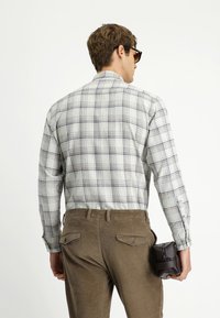 Mężczyzna w koszuli w kratę z długim rękawem i brązowych spodniach, trzymający małą ciemnobrązową skórzaną torbę, odwrócony plecami w stronę białego tła.