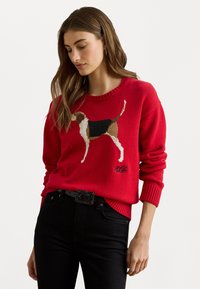 Rød genser med ribbestrikket hals og mansjetter, som har et brodert beagle-design i brunt, hvitt og svart, sammen med svarte jeans.