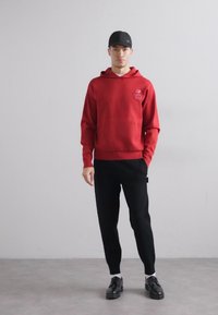 Felpa rossa con cappuccio, dotata di tasca frontale e dettagli del logo, abbinata a pantaloni neri e scarpe nere. Tessuto morbido con una vestibilità comoda.