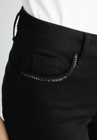 Pantalons noirs taille haute avec une texture lisse. Dispose d'une petite poche avant et de détails en sequins le long de l'ouverture de la poche.