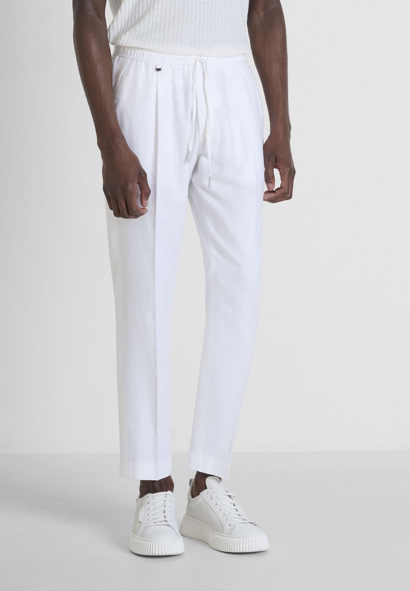 Homme portant un pantalon blanc à cordon avec un pli sur le devant et des baskets blanches, debout devant un fond clair uni.