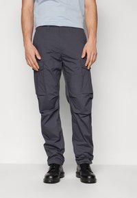 Pantalons cargo gris foncé en tissu léger, dotés de plusieurs poches latérales et d'une coupe décontractée. Portés avec des chaussures noires.