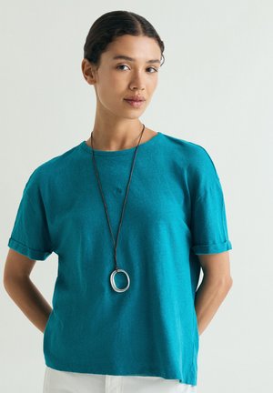 Joven mujer con cabello oscuro, camiseta turquesa y collar largo con colgante circular, de pie frente a un fondo claro liso.