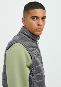 Chaleco de plumas gris con cuello alto y textura acolchada, usado sobre una camisa de manga larga verde. Presenta un cierre frontal y un diseño minimalista.