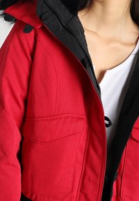 Červená a černá kapucí bunda s texturovanou látkou, zipovým zapínáním, dvěma předními kapsami a kapucí podšitou kožešinou.