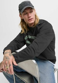 Sort sweatshirt med grøn broderi, denimjeans og en grå kasket med hævede bogstaver. Tatoverede hænder hvilende på en metalstol.