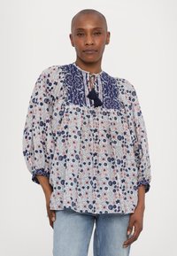 Weiße Bluse mit blauen Blumenmustern, ausgestattet mit einem marineblauen Kragen und einem Schlüssel-Loch-Bindeband. Lange Ärmel mit elastischen Bündchen und lockerer Passform.