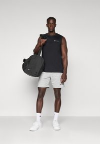 Black sleeveless shirt with a logo, gray shorts, white sneakers, and a black duffel bag. Plain background, standing pose.