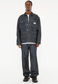 Donkere denimjas met contrasterende witte stiksels, voorzien van twee borstzakken en een label. Gecombineerd met bijpassende cropped donkere denimbroek.