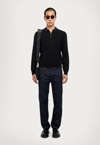 Polo shirt nero a maglia con zip e maniche lunghe, abbinato a jeans denim scuri a gamba dritta e scarpe nere. Il modello indossa occhiali da sole.