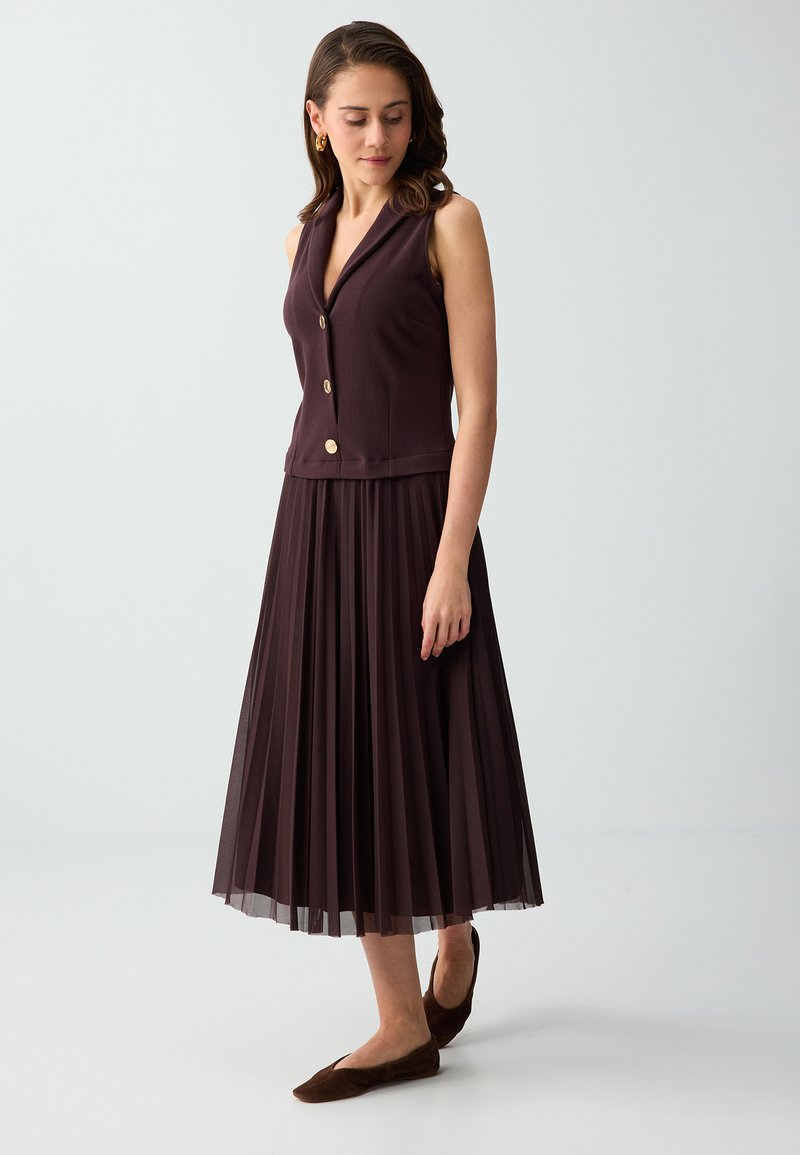 Femme portant un gilet sans manches violet foncé boutonné avec une jupe midi plissée et des chaussures plates assorties, debout sur un fond blanc.