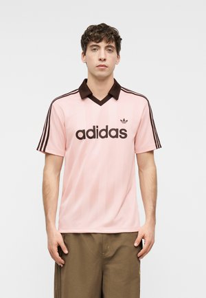 Joven de pie con una camiseta tipo polo Adidas de color rosa claro con cuello y rayas marrones, combinada con pantalones marrones.