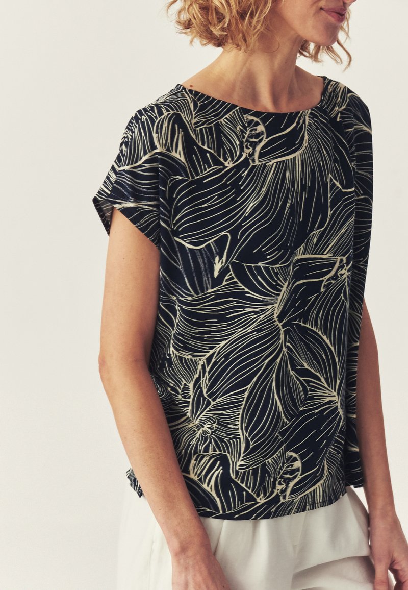 Femme portant un haut noir à manches courtes avec un grand motif floral blanc en ligne, associé à un pantalon de couleur claire, sur un fond uni.