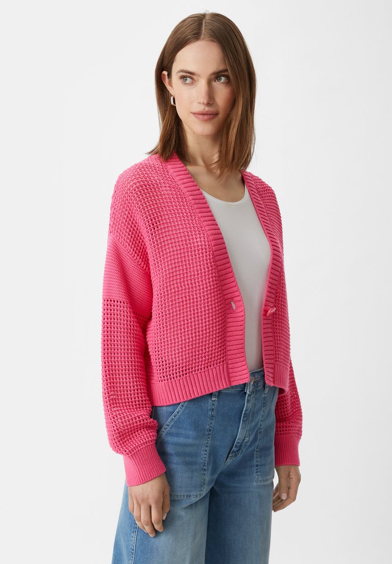 Różowy sweter z dzianiny ma fakturowany wzór, otwarty przód i ściągacze u rękawów. Nosi się go na białym topie z luźnymi niebieskimi dżinsami.