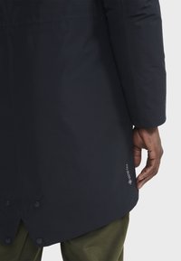 Veste imperméable bleu foncé avec une texture lisse, boutons en bas et logo Gore-Tex près de l'ourlet. Une main tient le bord de la veste.