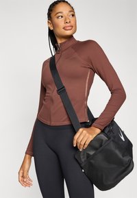 Καφέ μακρυμάνικο μπουφάν με φερμουάρ μπροστά, συνδυασμένο με μαύρα κολάν και μαύρη τσάντα με δίχτυ και λευκό λογότυπο. Λεία υφή.